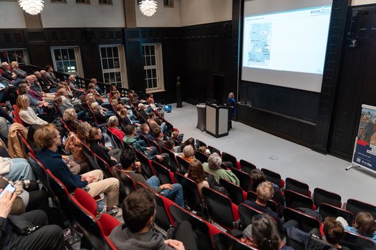 Publikum bei Infoveranstaltung Das Foto zeigt den fast voll besetzten Hörsaal des BNITM. Vorne steht eine Rednerin am Pult, im Hintergrund sieht man eine Folie der Präsentation.