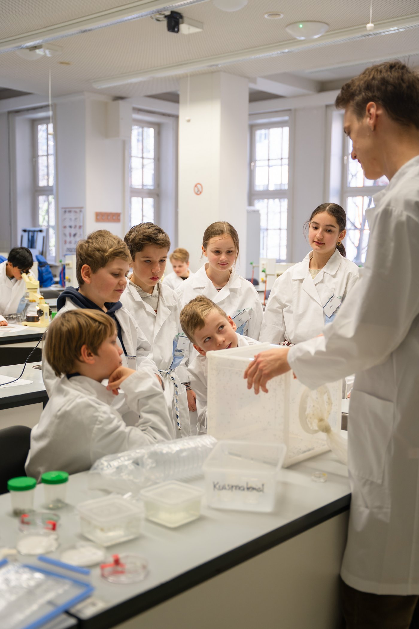 Eine Gruppe von sechs Schüler:innen steht im Halbkreis vor einem Kasten, in dem sich Mücken befinden. Ein Forscher erklärt ihnen die Mückenforschung. Alle tragen weiße Kittel.