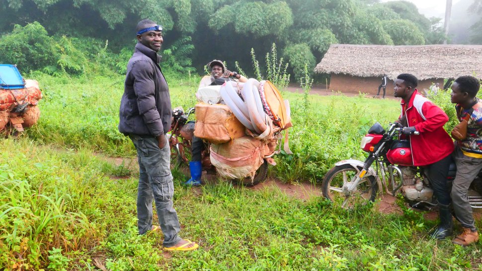 Reisende Händler in der DRC Auf dem Foto sind zwei Motorräder mit einer bzw. zwei Personen darauf zu sehen. Im Vordergrund steht ein Mann dunkler Kleidung. Sie befinden sich draußen im Grünen.