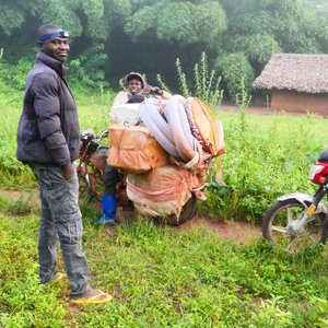 Reisende Händler in der DRC Auf dem Foto sind zwei Motorräder mit einer bzw. zwei Personen darauf zu sehen. Im Vordergrund steht ein Mann dunkler Kleidung. Sie befinden sich draußen im Grünen.