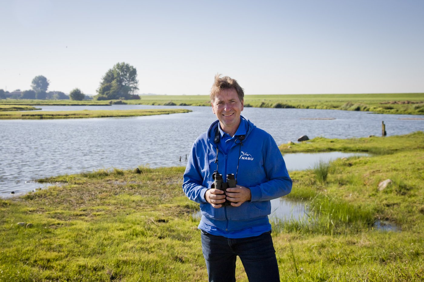 Das Foto zeigt einen Mann mittleren Alters mit Fernglas in der Hand vor einer Seenlandschaft stehen und in die Kamera lächeln.