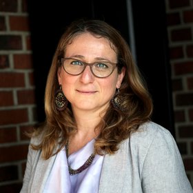 Daniela Fusco, Ph.D.: a researcher against a dark red brick background, wearing her blonde shoulder-length hair loose.