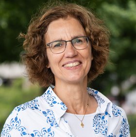 Prof. Iris Bruchhaus, a researcher with a white and blue blouse, curly mid-length hair, glasses and an open smile