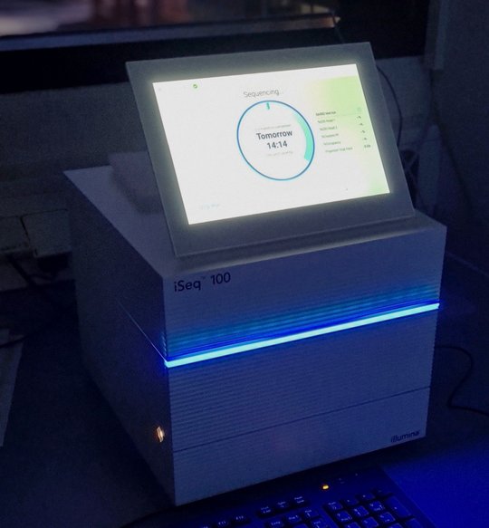 iSeq100 Sequencer Zu sehen ist eine Aufnahme in einem verdunkelten Raum. Ein weißes Gerät mit einem großen Monitor erhellt die Dunkelheit. Der Monitor leuchtet in weiß, zu sehen ist ein Kreis, in dem Tomorrow 14:14 steht. an dem Gerät leuchtet außerdem eine kleine blaue Leiste.