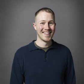 A smiling man with short hair is wearing a dark blue collared shirt against a gray background.