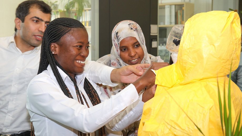 The photo shows three diverse people helping a fourth to put on a personal protective suit.