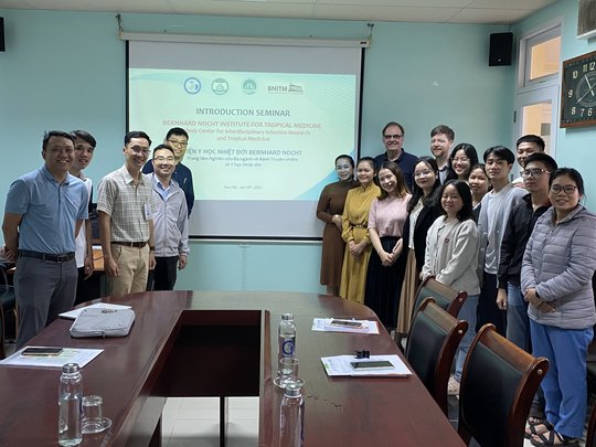 The picture shows students and lecturers from the Institute for Community Health Research and the Faculty of Public Health as well as Dr. Jörg Blessmann and Dr. Benno Kreuels after the presentation. The picture shows several people standing in front of a projected presentation. They are all smiling at the camera.