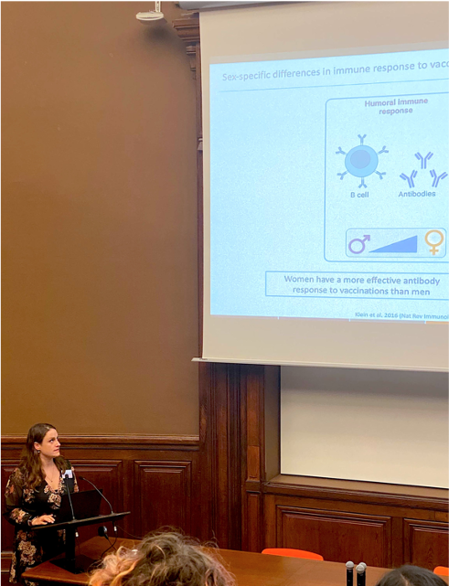Dr. Charlotte Hansen The picture shows Charlotte Hansen in a lecture hall at the Pasteur Institute. She is small in the bottom left of the picture, while her presentation is large in the background. Charlotte is wearing a dark dress with flowers.